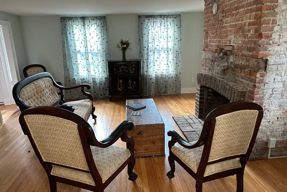 A cozy living room featuring vintage chairs, a wooden coffee table, a brick fireplace, and sheer curtains.