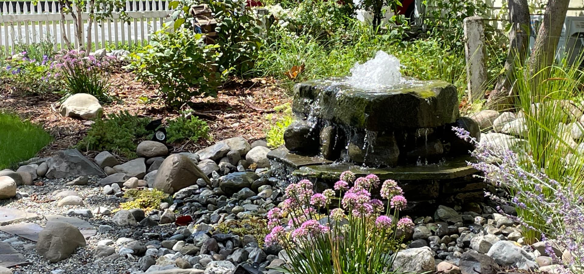 A serene garden featuring a small stone fountain surrounded by colorful flowers and pebbles.